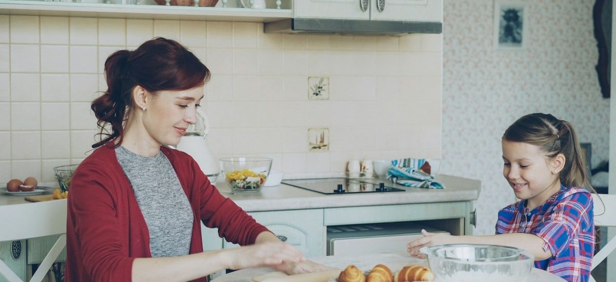 Young mother rolling dough and talking to her little cute daughter while cooking in the kitchen on weekend. Family, food, home and people concept